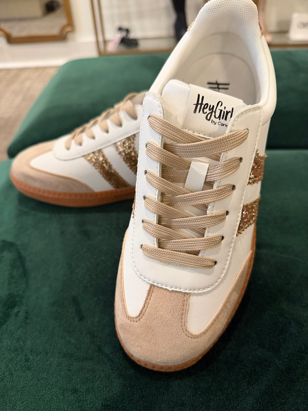 A pair of low-top sneakers by HeyGirl by Casm, featuring off-white leather, beige suede panels, beige laces, and gold glittery side stripes, displayed on a dark green velvet surface.