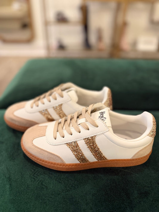 A pair of white low-top sneakers with beige suede overlays, gold glitter three stripes, beige laces, and gum soles, displayed on a dark green velvet surface.